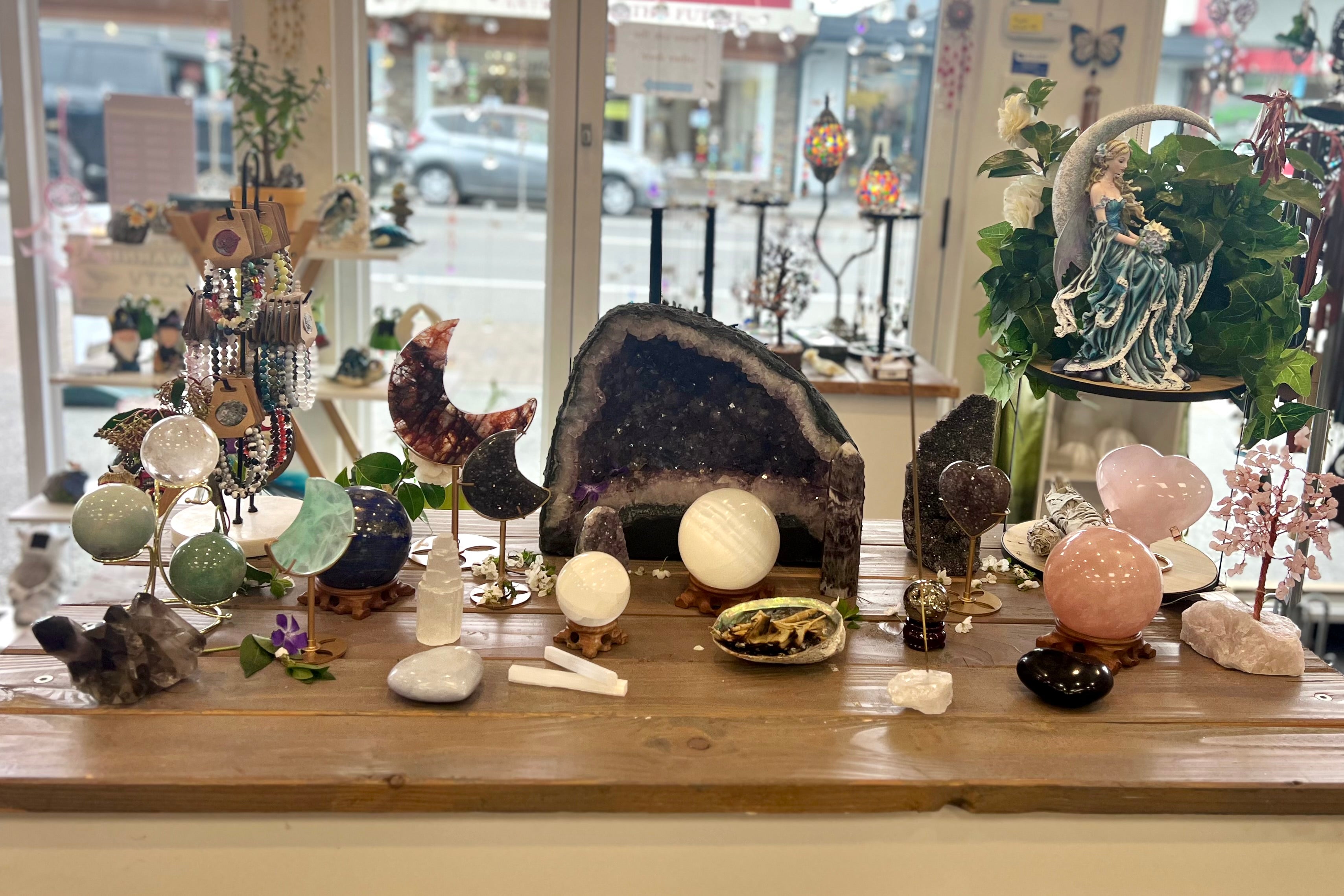 A window display of crystals in the Simplyb shop in Kaiapoi: a large amethyst geode, polished spheres, selenite, rose quartz and a smudge bundle on a timber table, with Williams Street visible through the glass behind