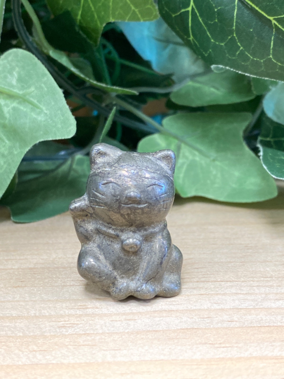 Small grey pyrite lucky cat figurine sitting on pale wood with green potted plants behind
