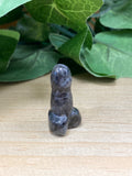 Labradorite mini penis carving, dark grey-blue polished stone, standing upright on wooden surface with green leaves behind