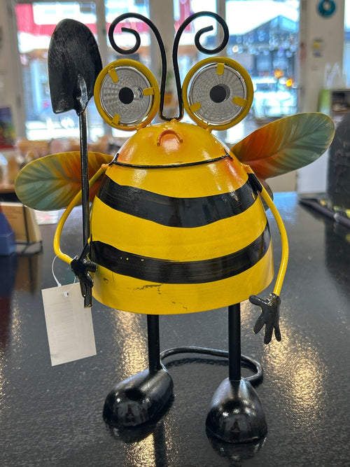 Yellow and black metal bee garden ornament with reflector eyes, shovel, and green wings, indoors