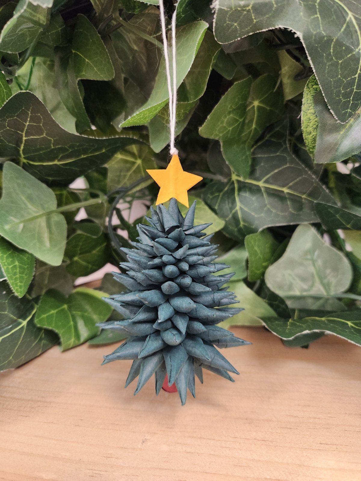Blue stone Christmas tree ornament with yellow star topper, hanging in front of green foliage.