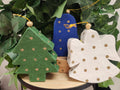 Three fabric Christmas ornaments with gold polka dots: green tree, blue figure, white angel, nestled in fresh foliage.