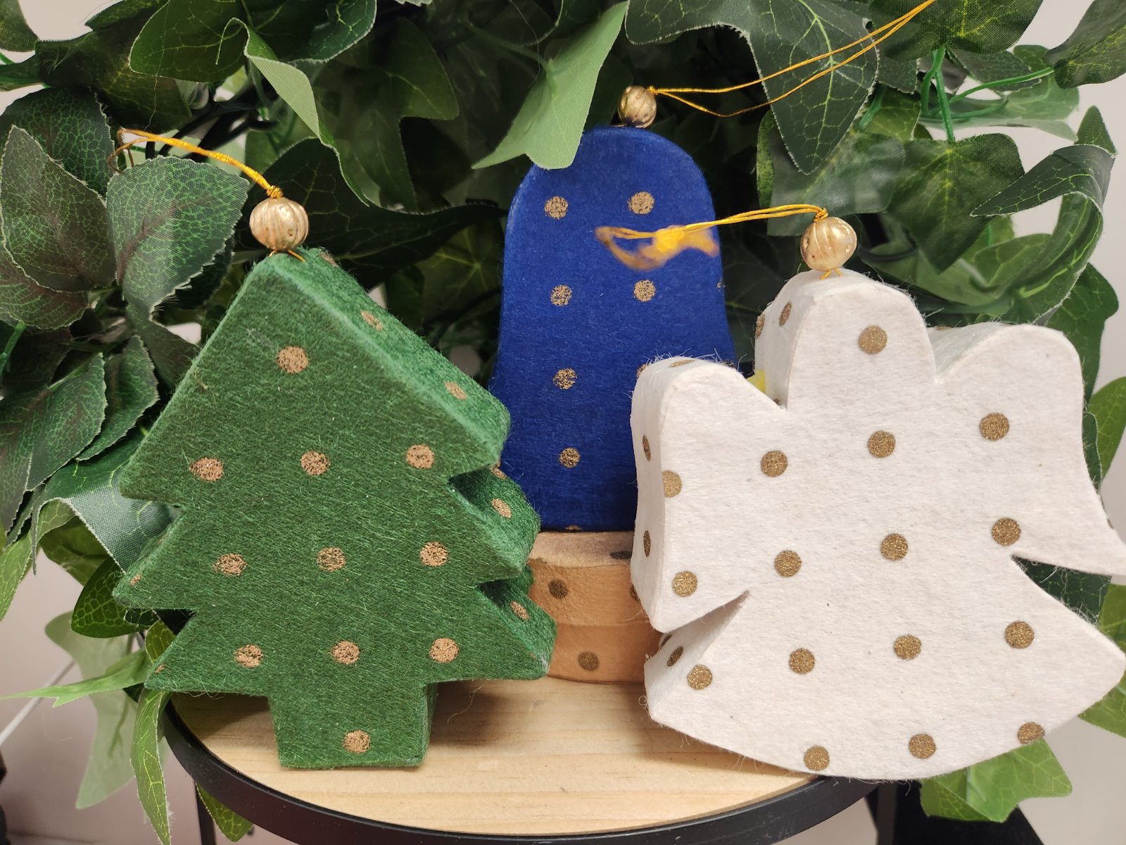 Three fabric Christmas ornaments with gold polka dots: green tree, blue figure, white angel, nestled in fresh foliage.