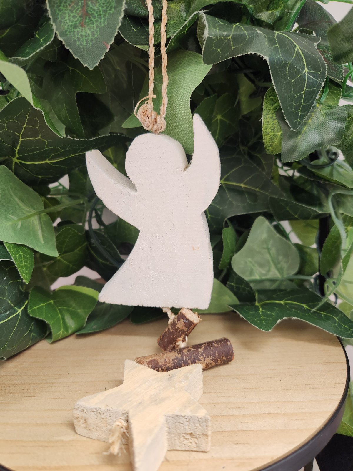 White ceramic hanging angel ornament with crescent moon shape, suspended by twine above wooden stand with small branches.