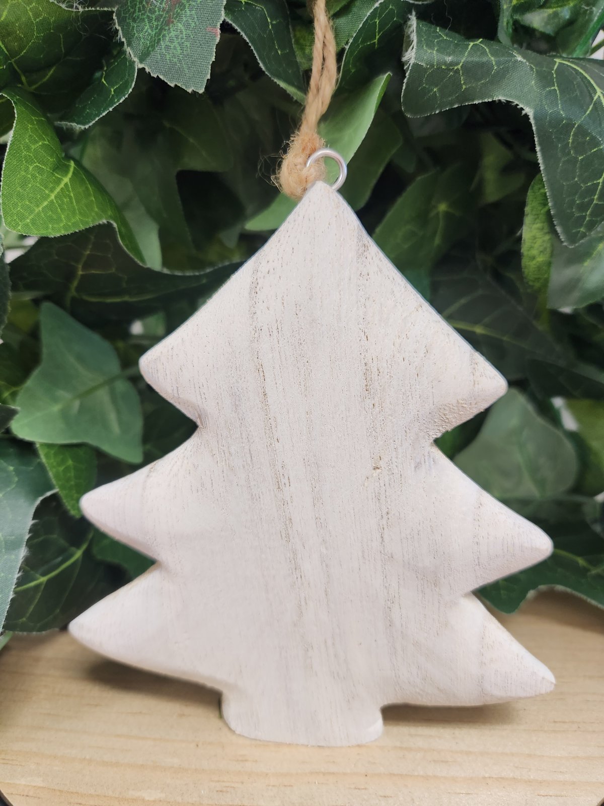 White washed wooden Christmas tree ornament with twine loop, hanging in front of green leaves