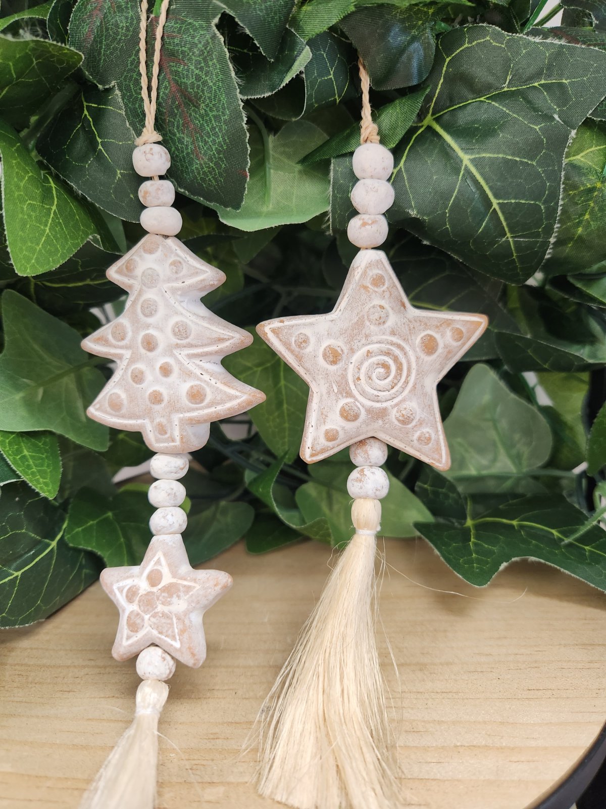 Two wooden Christmas ornaments hanging from twine: a tree with dot pattern and a star with spiral detail, strung with beads and tassels.