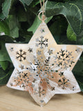 Wooden star ornament with laser-cut snowflakes and stars, natural finish with string hanger, surrounded by green foliage