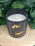 Black glass candle with gold celestial design, white wax visible, surrounded by green foliage