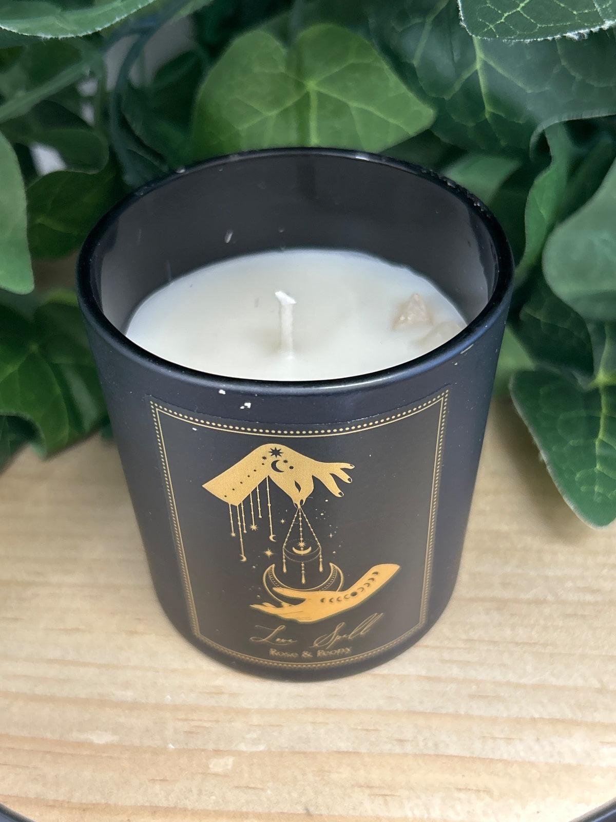 Black glass candle with gold celestial design, white wax visible, surrounded by green foliage