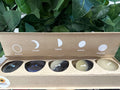 Five scented candles in kraft box with moon phase symbols and labels: Geranium, Lavender, Cypress, Jasmine, Bergamot.