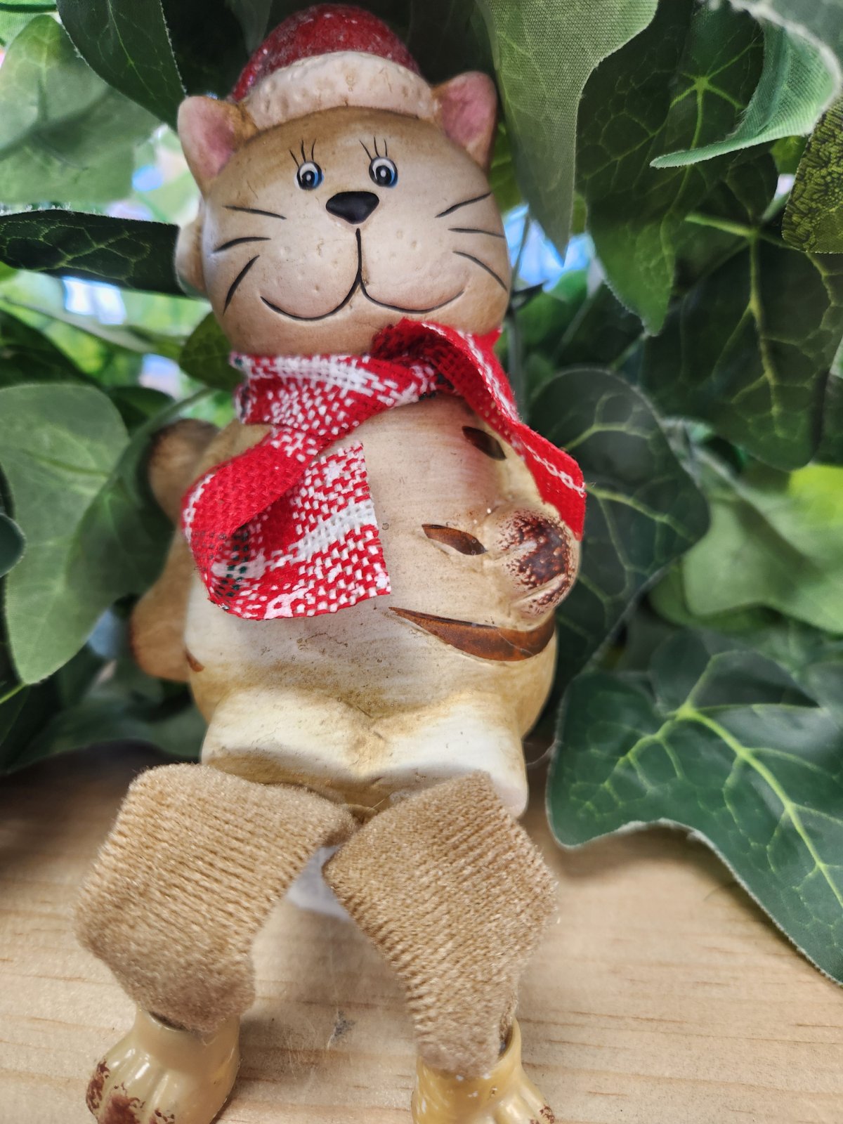 Ceramic cat figurine in Santa hat and red-and-white scarf, with swing legs and a warm smile, sitting among green leaves.