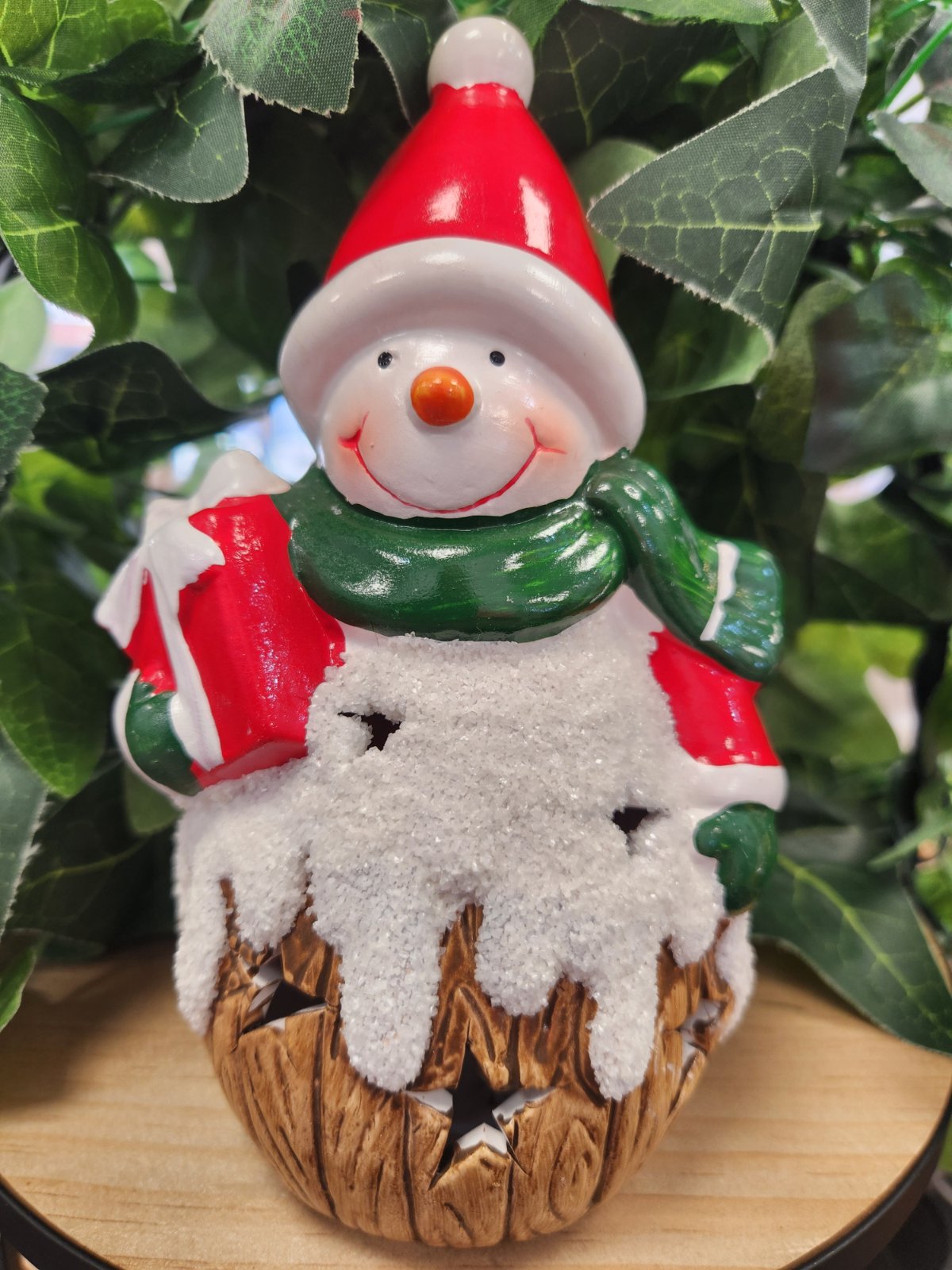Ceramic snowman ornament with red hat, green scarf, and textured base, holding a red object.