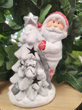 White ceramic Santa ornament holding a silver star, dressed in red and white, displayed among green leafy branches