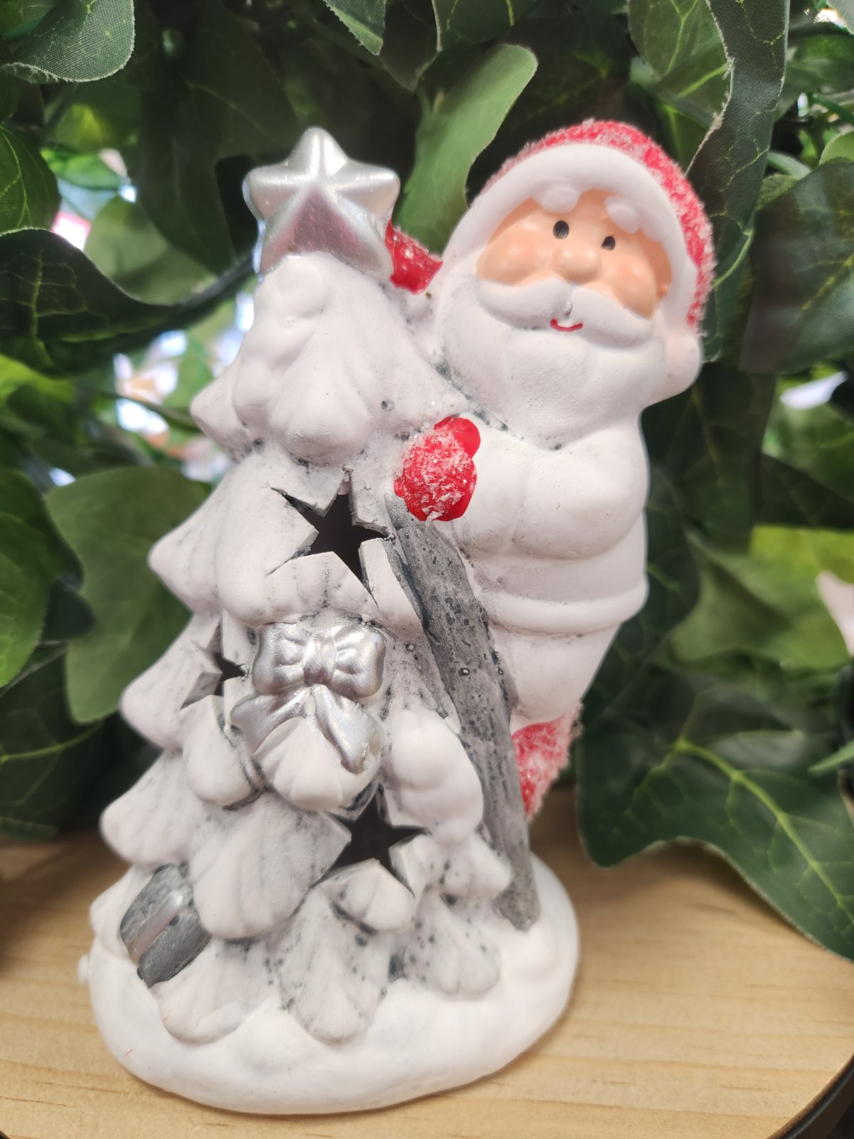 White ceramic Santa ornament holding a silver star, dressed in red and white, displayed among green leafy branches
