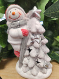 White ceramic snowman figure hugging a frosted evergreen tree with red and silver glitter accents and a bow