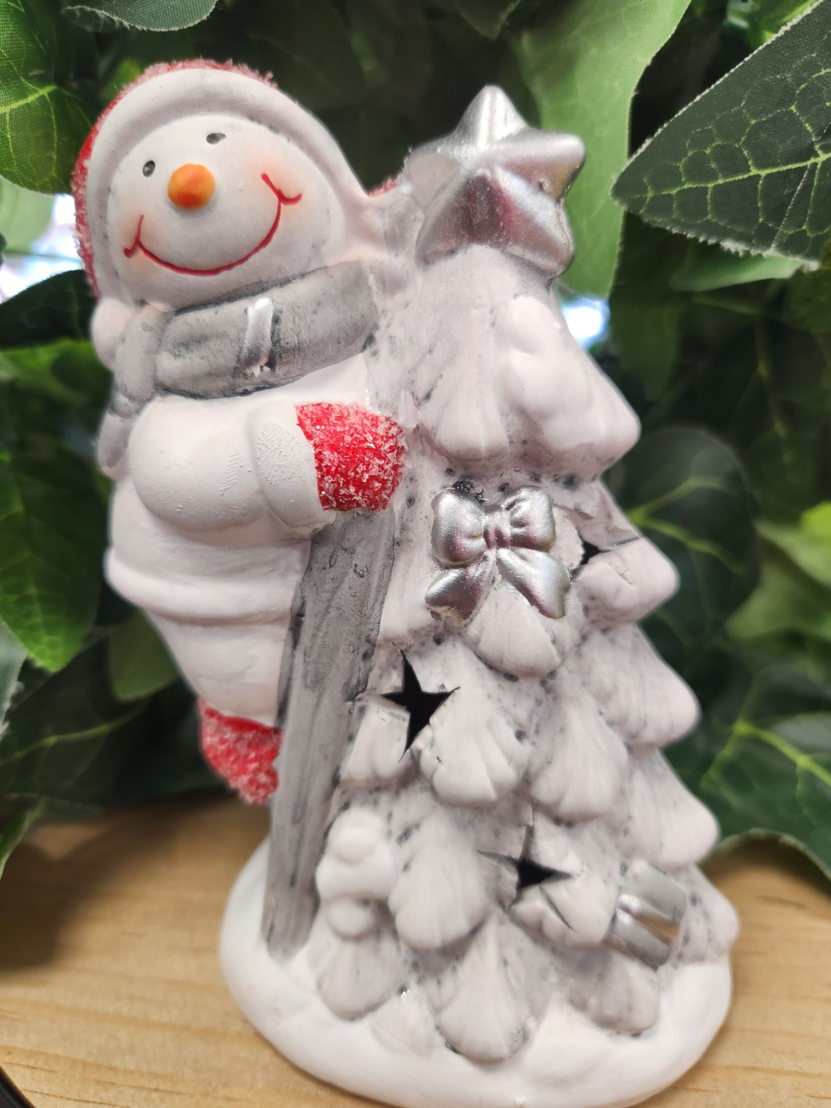White ceramic snowman figure hugging a frosted evergreen tree with red and silver glitter accents and a bow