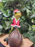 Christmas frog figurine in red Santa suit with white trim, holding a dark brown sack, perched on a branch among green leaves.