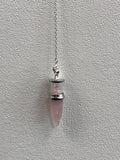 Rose quartz bullet pendulum hanging from a silver chain against a textured white wall