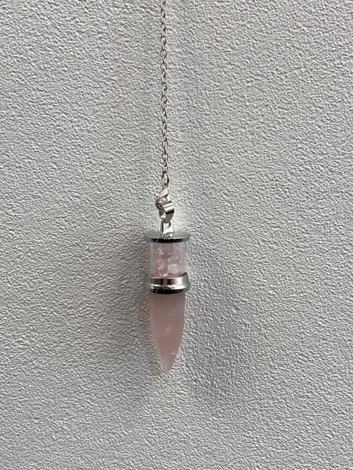 Rose quartz bullet pendulum hanging from a silver chain against a textured white wall