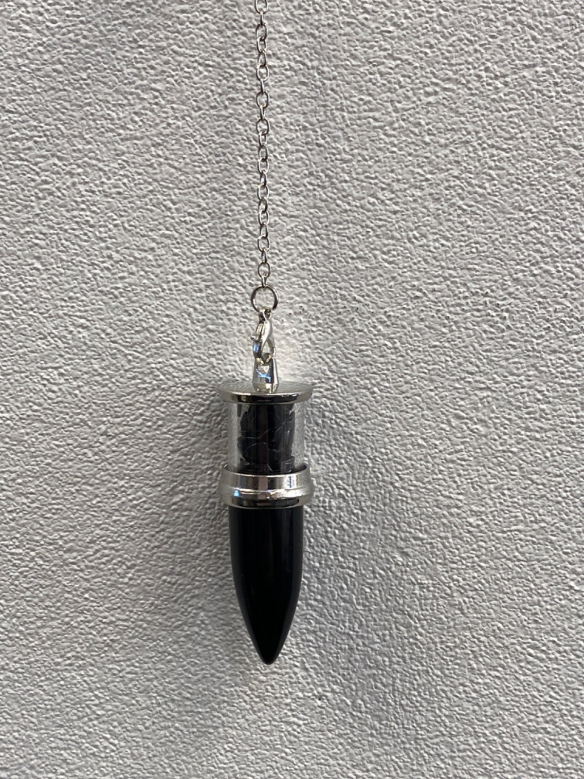 Black onyx bullet pendulum hanging from a silver chain against a textured white wall