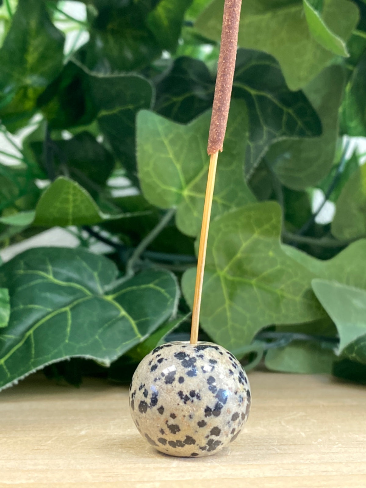 Dalmatian Jasper sphere incense holder with speckled cream and dark markings on wooden surface, green foliage behind