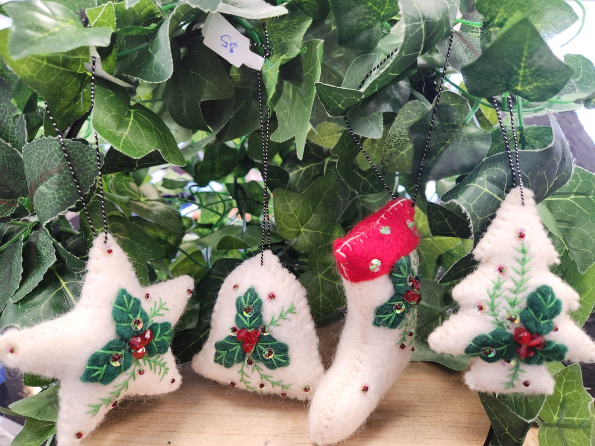 Five white felt Christmas ornaments with embroidered holly leaves and red berries, hanging from chains among green foliage