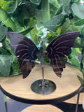Dark obsidian butterfly ornament on metal stand, displayed among green leafy plants on wooden surface.