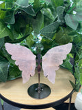 Rose quartz crystal butterfly with translucent pale pink wings and black metal body on a display stand, surrounded by green foliage.