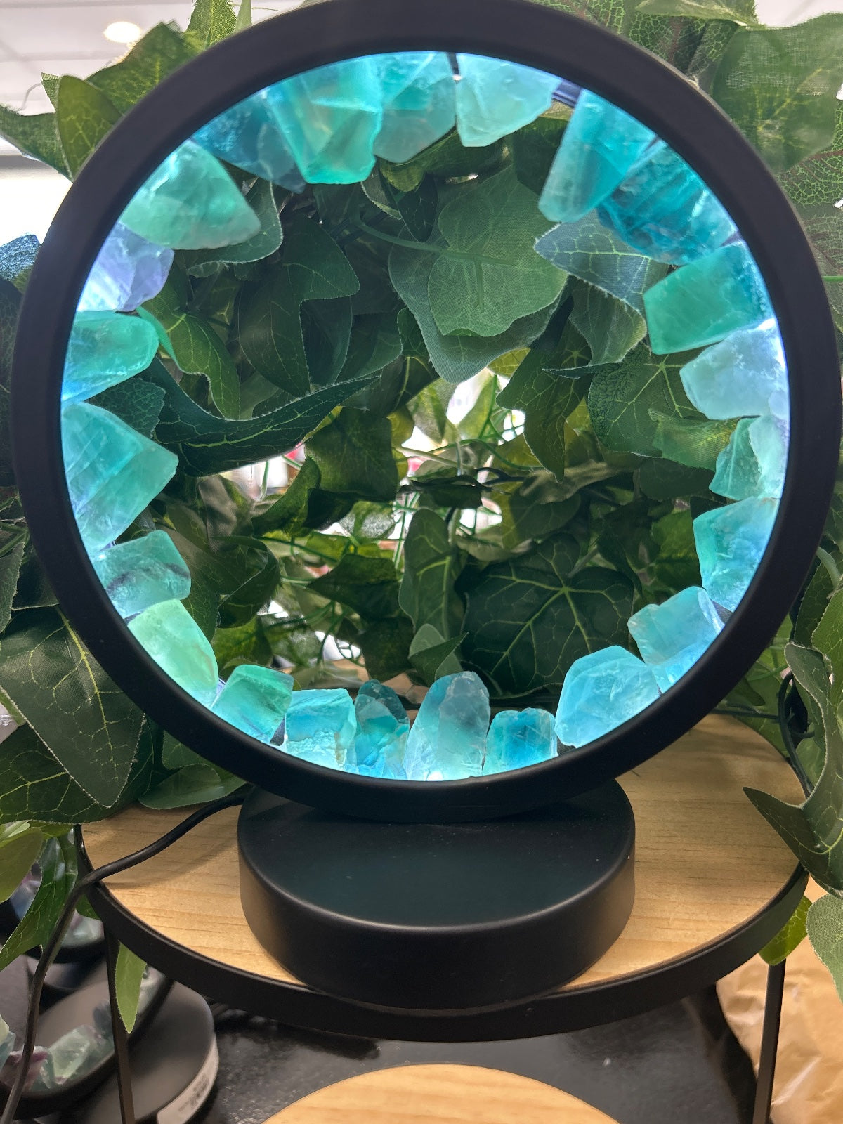 Black circular LED lamp with turquoise glow, surrounded by green foliage and climbing ivy plants