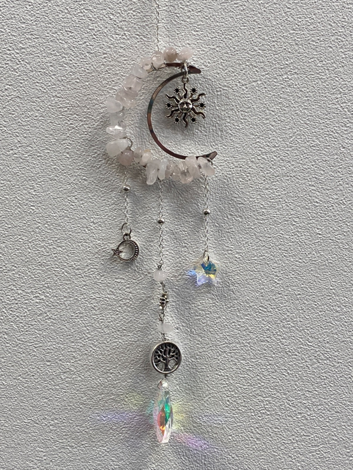 Rose quartz chip crescent moon suncatcher with dangling charms against white textured wall.