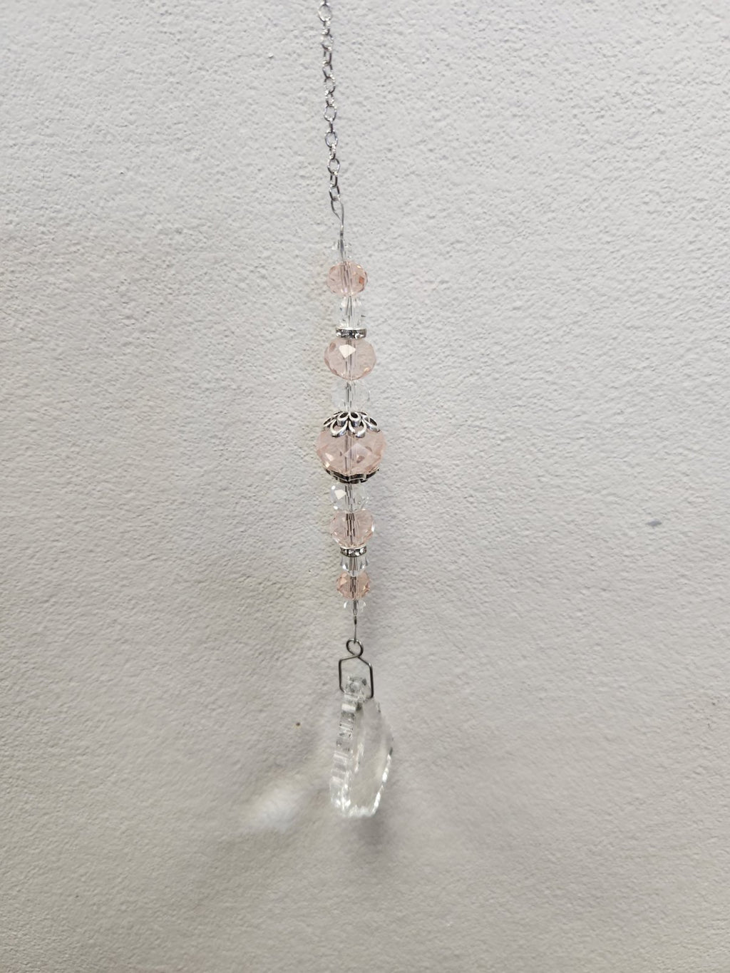 Pink beaded suncatcher with silver chain, decorative caps, and clear crystal teardrop pendant hanging against white wall