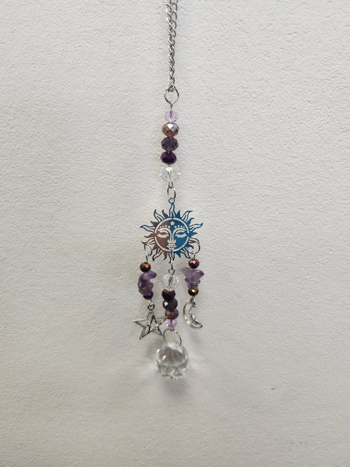 Silver chain hanger with purple amethyst beads, a blue and copper sun-moon face charm, and dangling star and crescent charms.