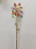 Metal stake flower with faceted amber and multicoloured crystal petals, green beads along the stem, against white wall