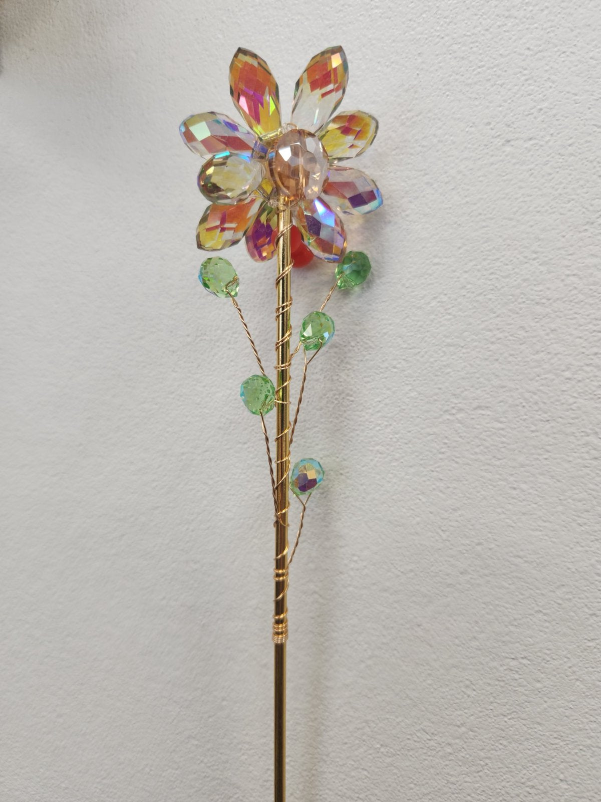 Metal stake flower with faceted amber and multicoloured crystal petals, green beads along the stem, against white wall