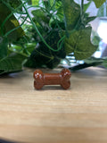 Mahogany Jasper bone carving in warm reddish-brown, shaped like a dumbbell with rounded bulbs at each end, on wooden surface with green plant behind.