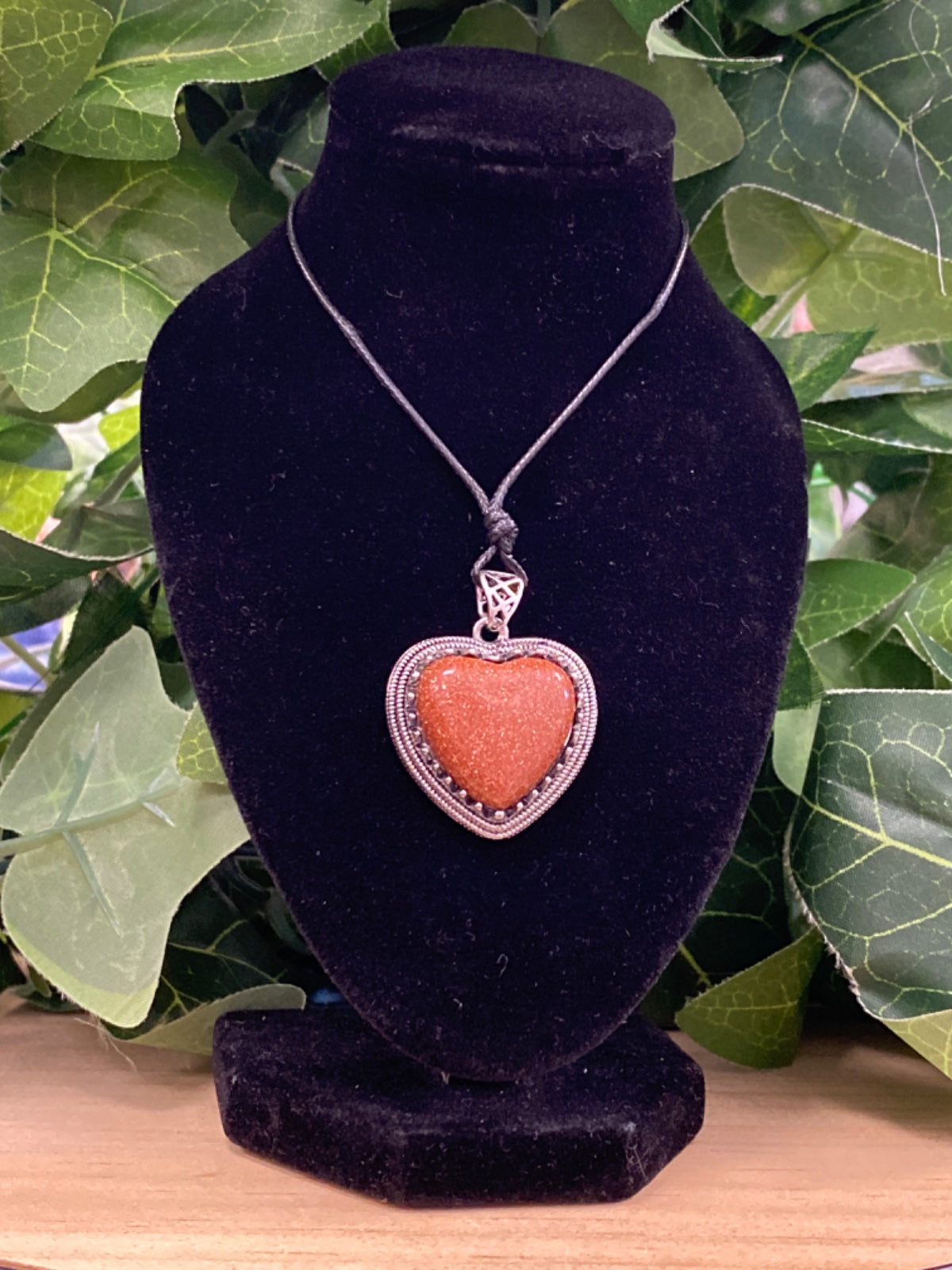 Red goldstone heart pendant on silver chain, displayed on black velvet bust with green foliage behind