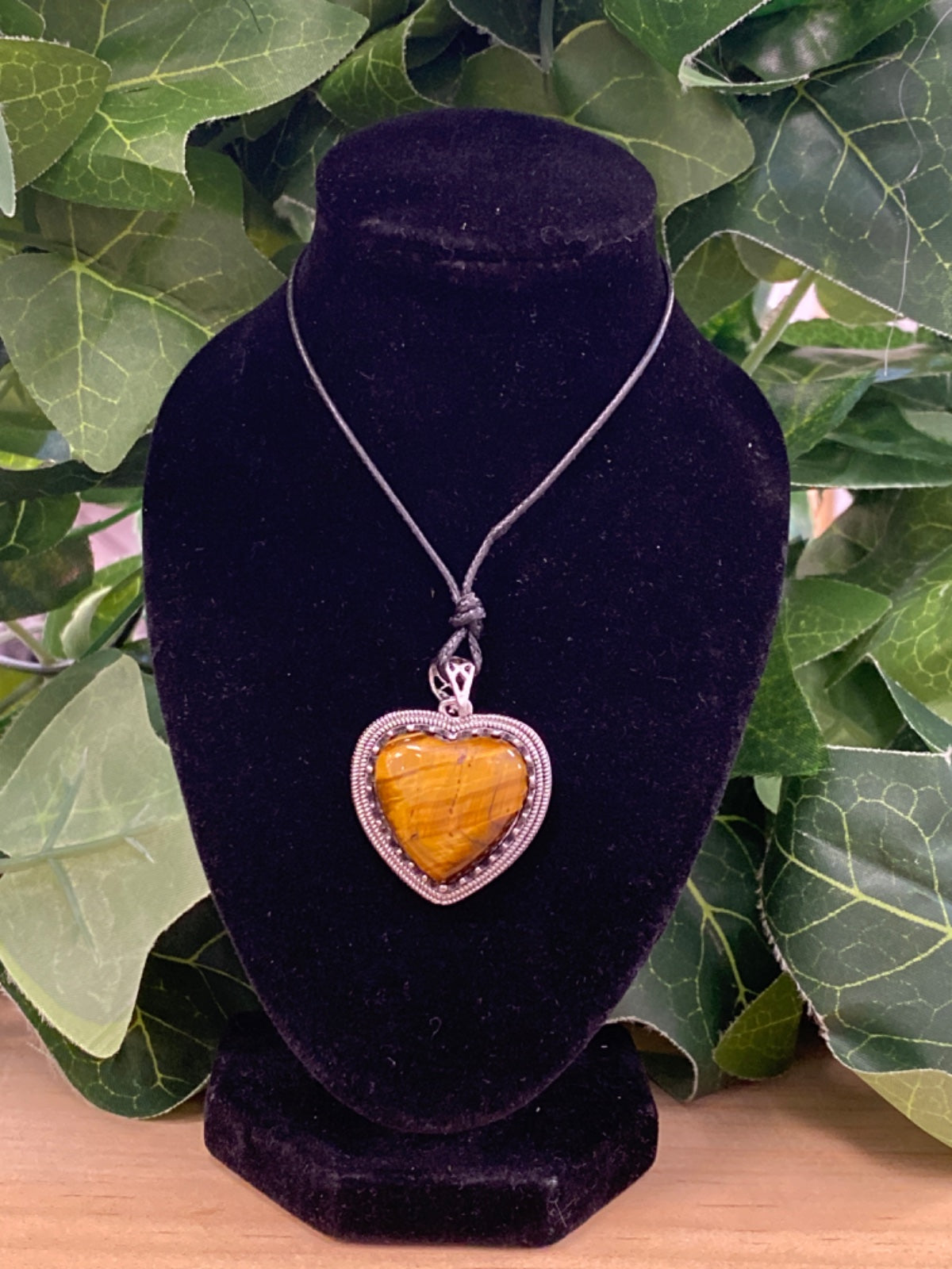 Tigers eye heart pendant in silver bezel on black cord, displayed on black velvet bust with green foliage backdrop