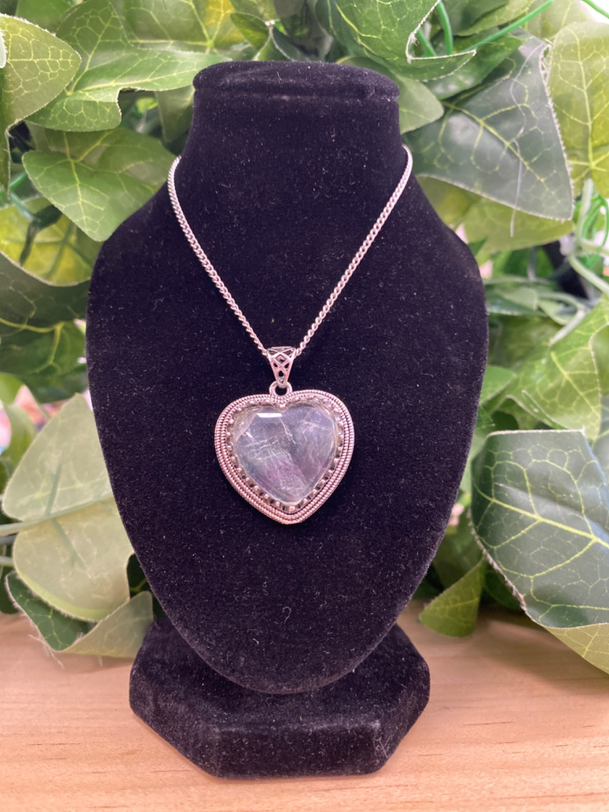 Fluorite heart pendant on silver chain displayed on a black velvet bust against green foliage