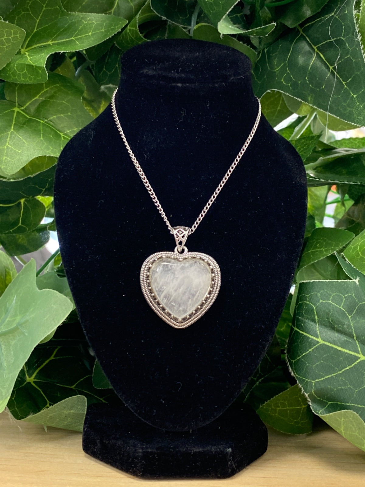 Clear quartz heart pendant on silver chain displayed on black velvet necklace bust with green leaves behind