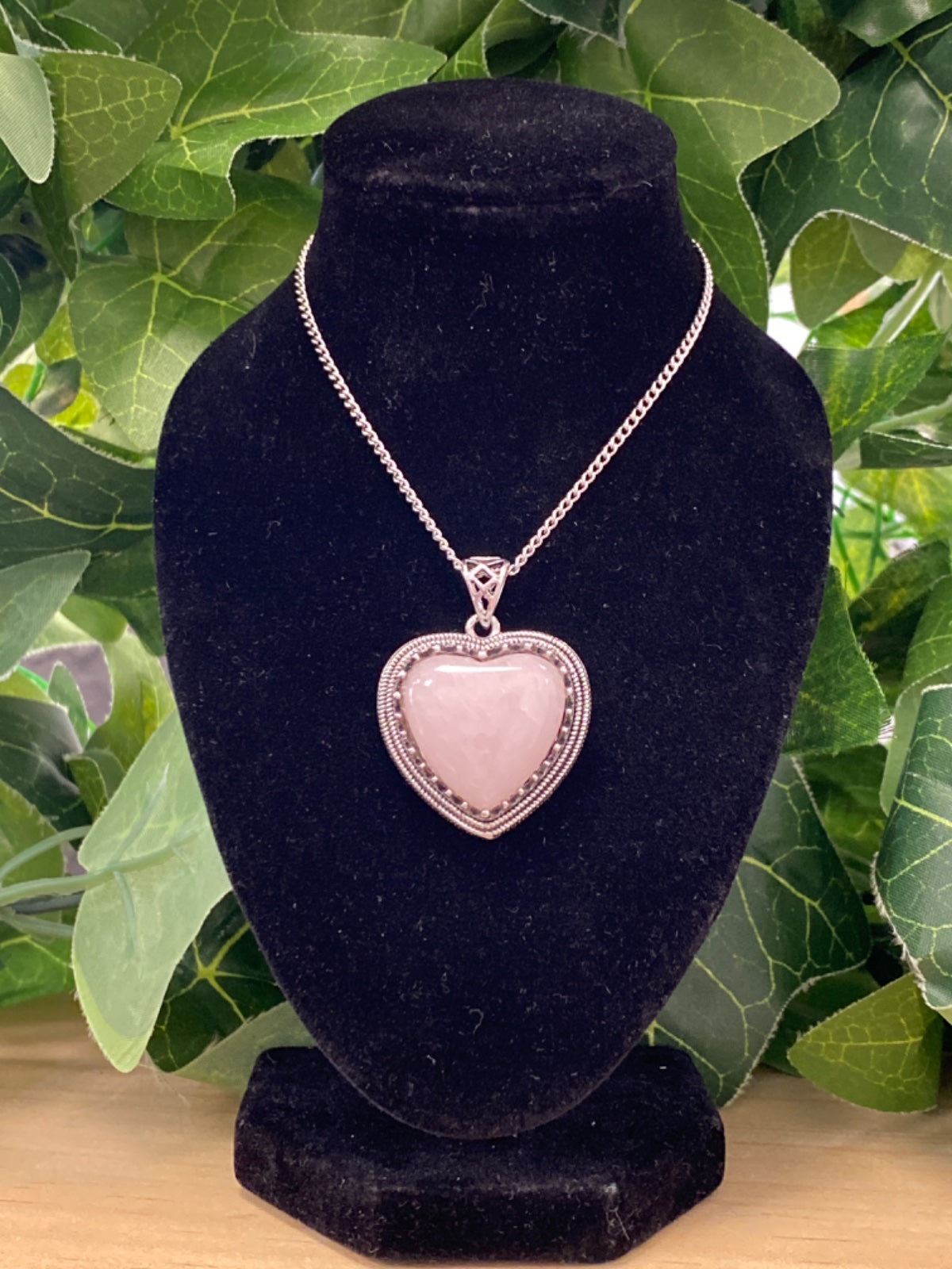 Rose quartz heart pendant on silver chain, displayed on black velvet bust with green foliage background