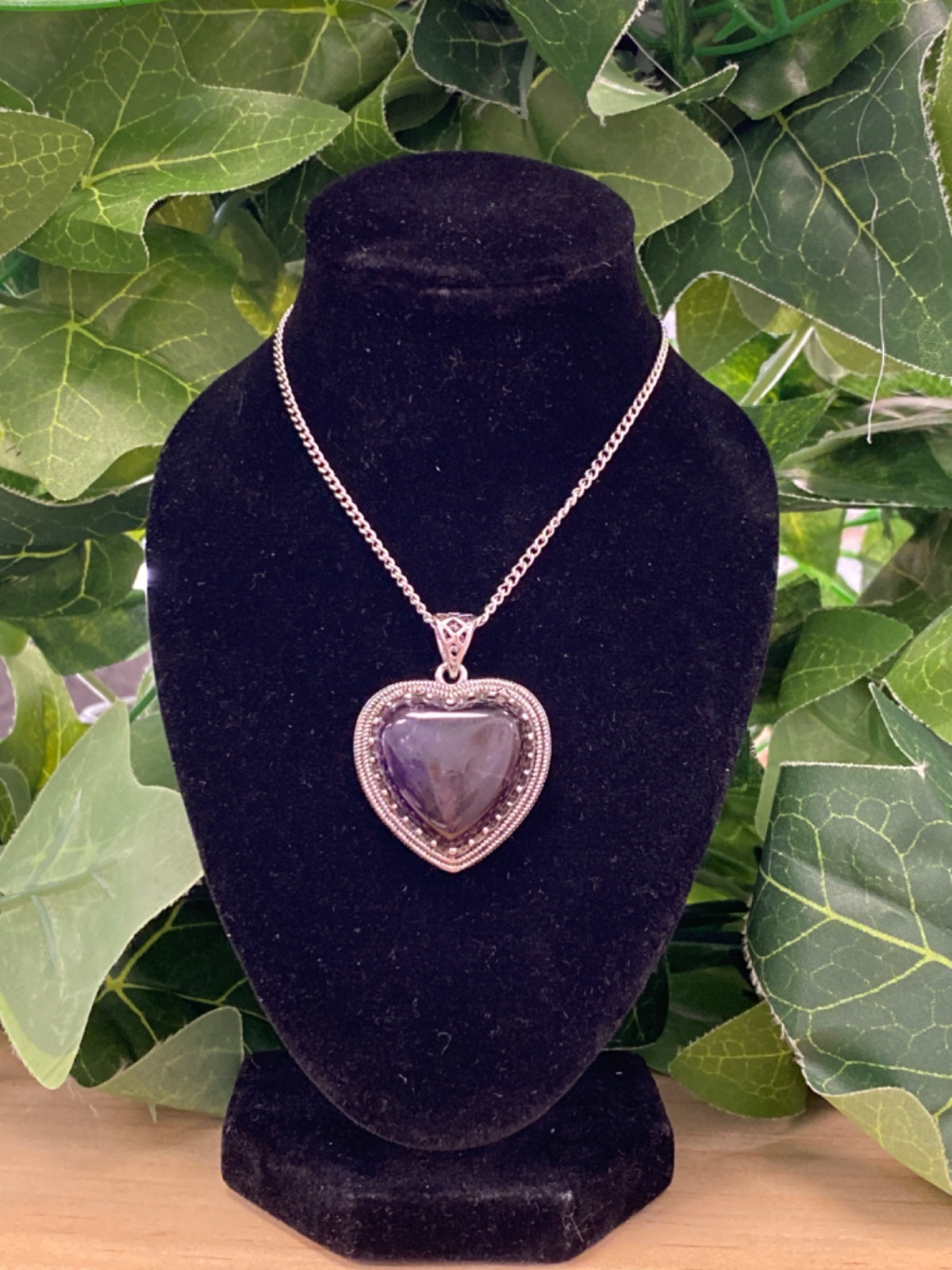 Amethyst heart pendant on silver chain displayed on black velvet bust stand, surrounded by green foliage