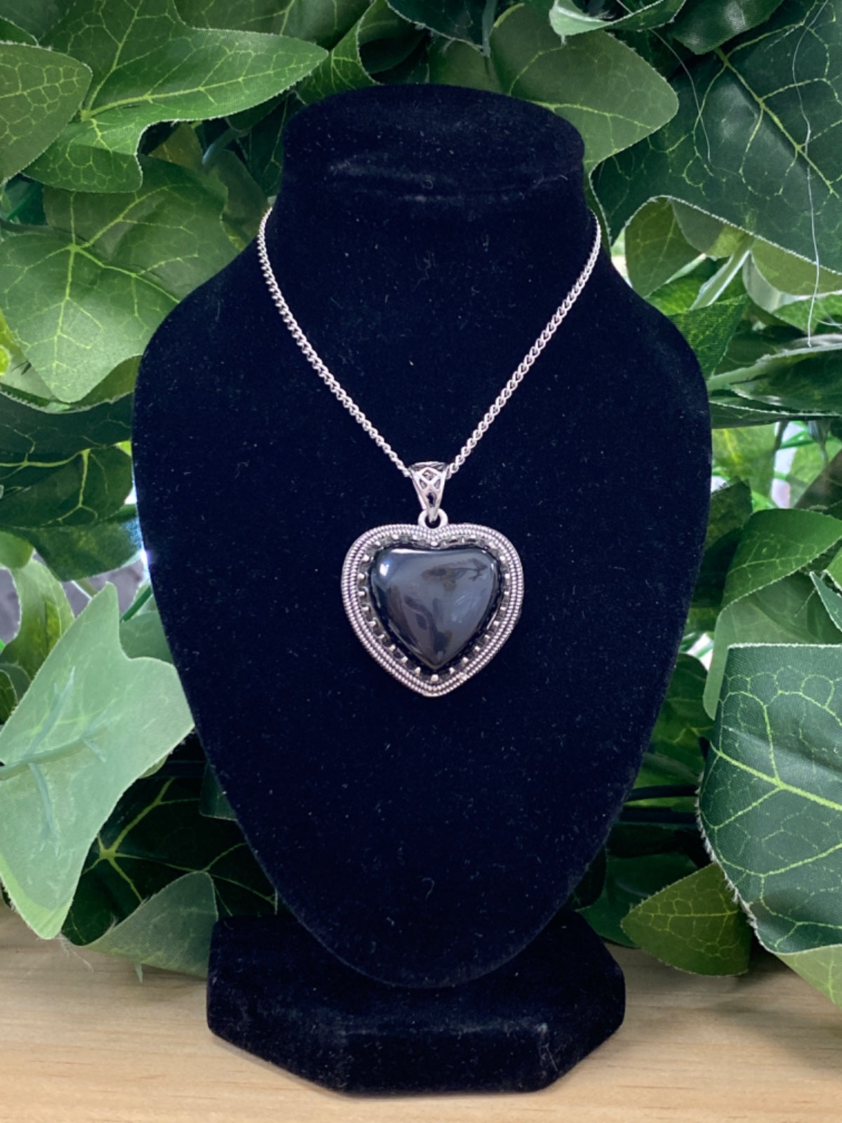 Black agate heart pendant with silver beaded frame on silver chain, displayed on black velvet bust with green foliage behind