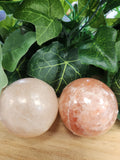 Two polished Himalayan salt massage balls, one pale cream and one warm peachy-pink, nestled among green leaves.
