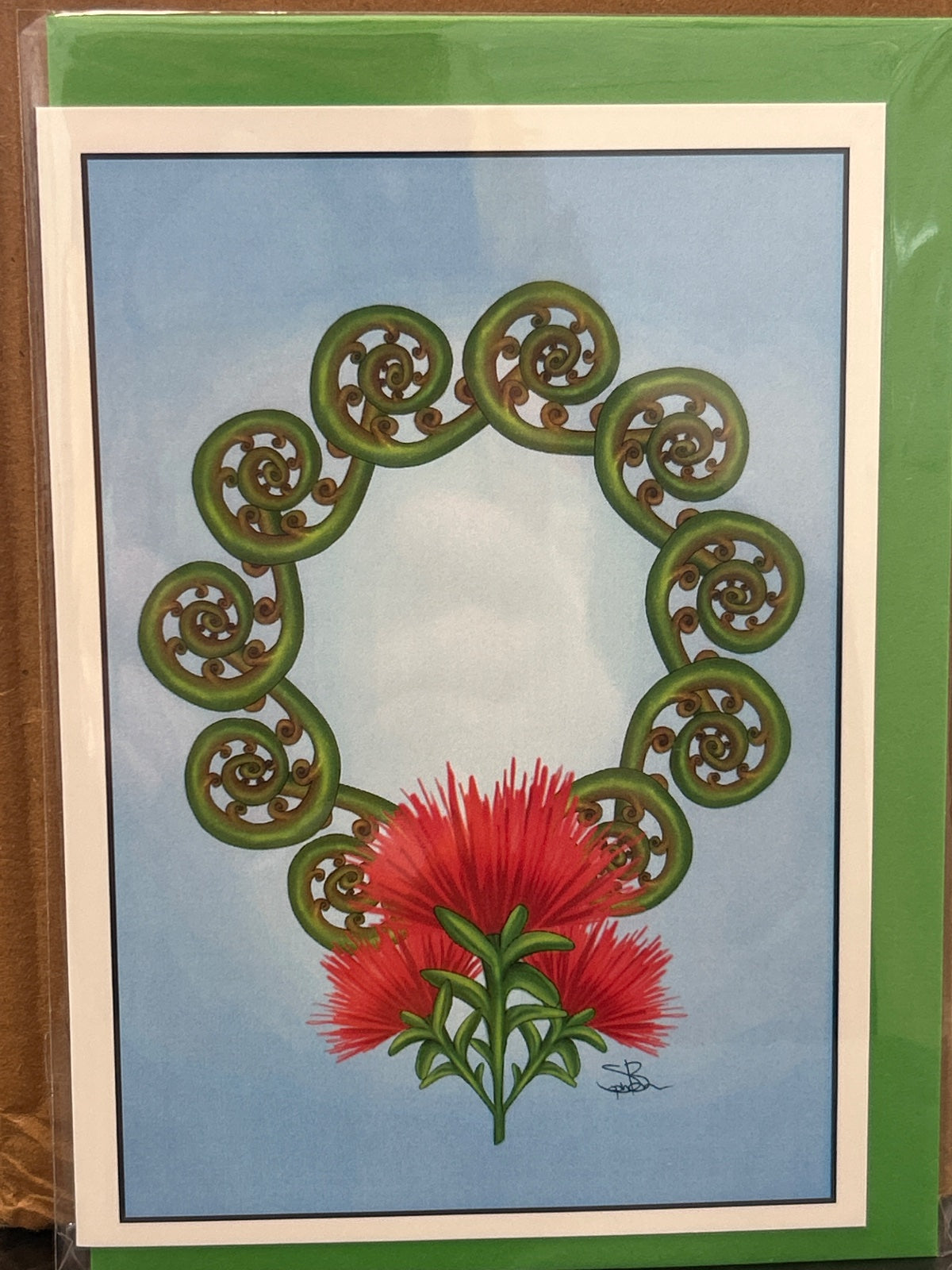 Greeting card with ten green spiral koru arranged in a circle around a bright red bottlebrush flower on a light blue background.