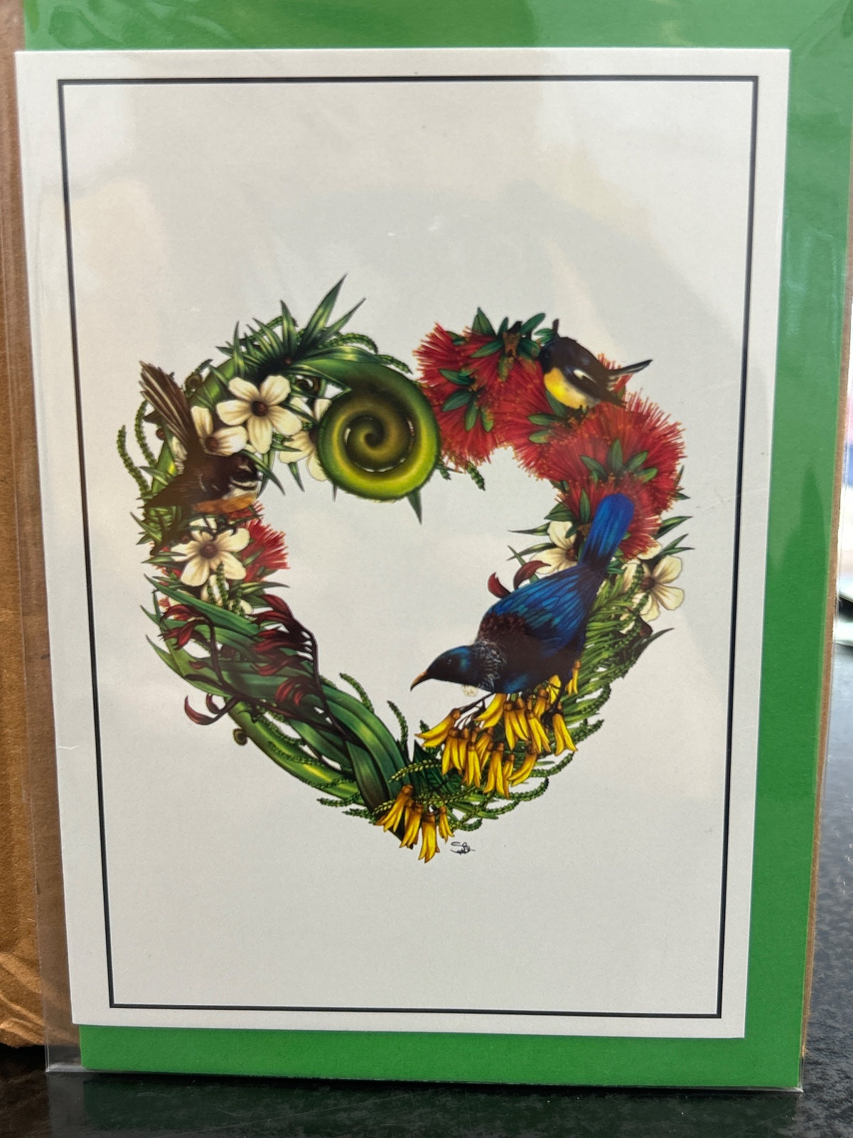 Heart-shaped greeting card with NZ native bird and flowers including red bottlebrush, white blossoms, yellow flowers, and a blue tui bird