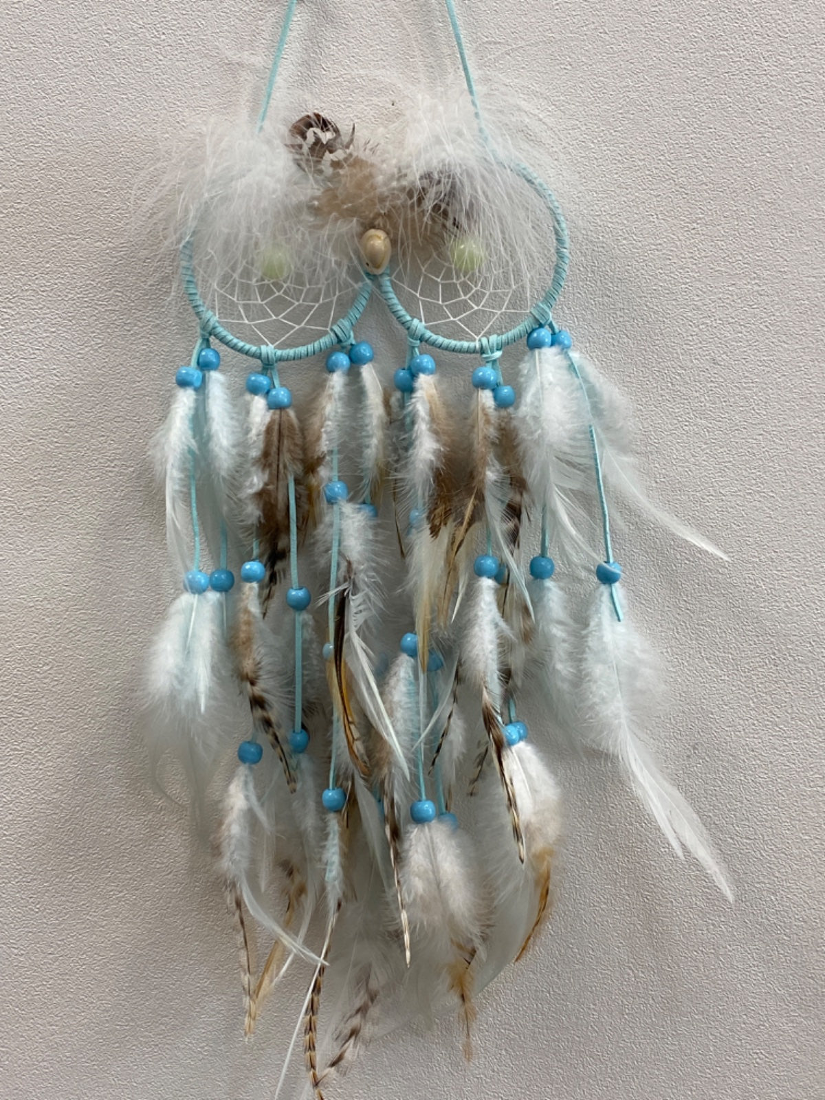 Blue and brown feather dream catcher with turquoise web, beaded strands, and natural feathers hanging against white wall.