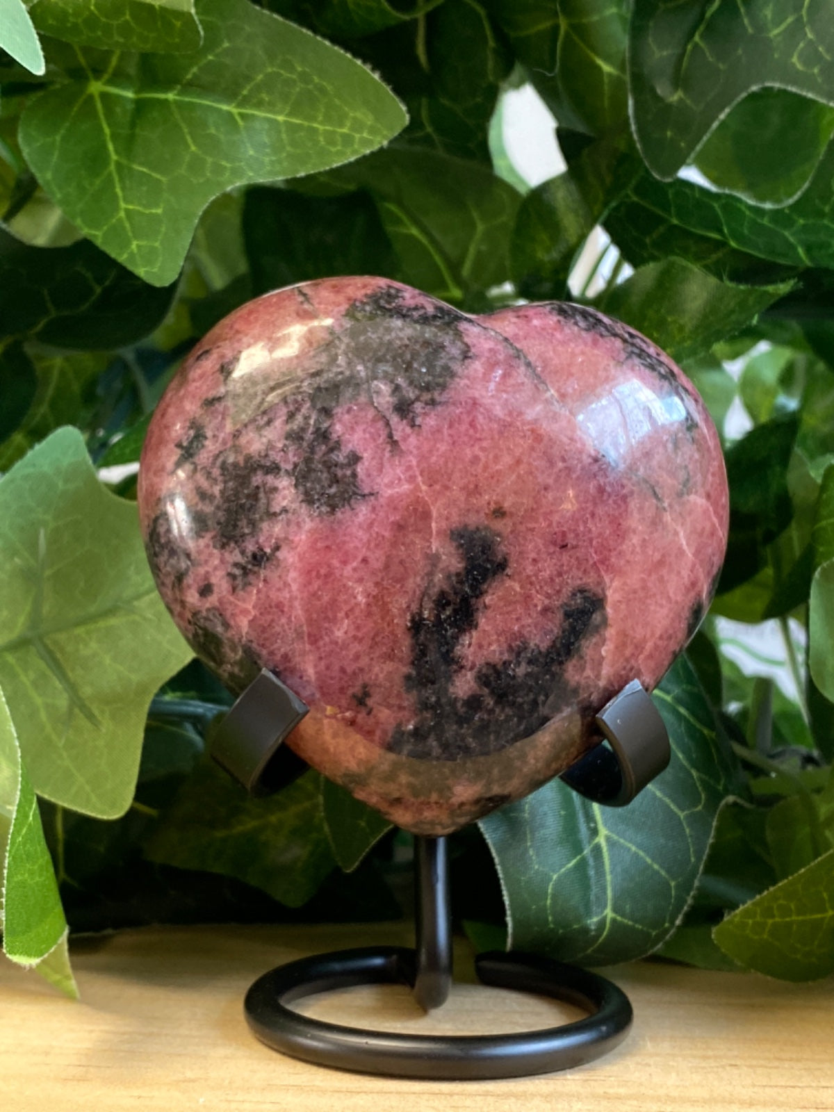 Polished rhodonite heart, 6cm, dusty pink with black manganese oxide markings, on a black metal stand surrounded by green foliage.