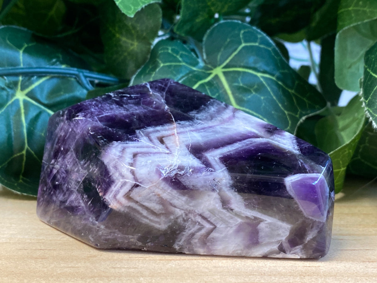 Purple and white banded amethyst chevron freeform, 223g, on wooden surface with green foliage behind