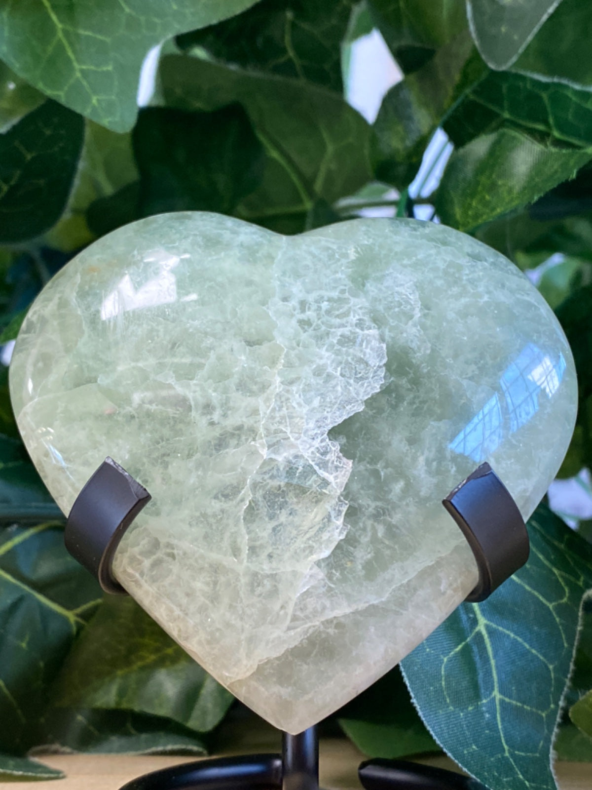 Pale green fluorite heart, 6.5cm, polished with natural fracture patterns, mounted on black stand with foliage behind