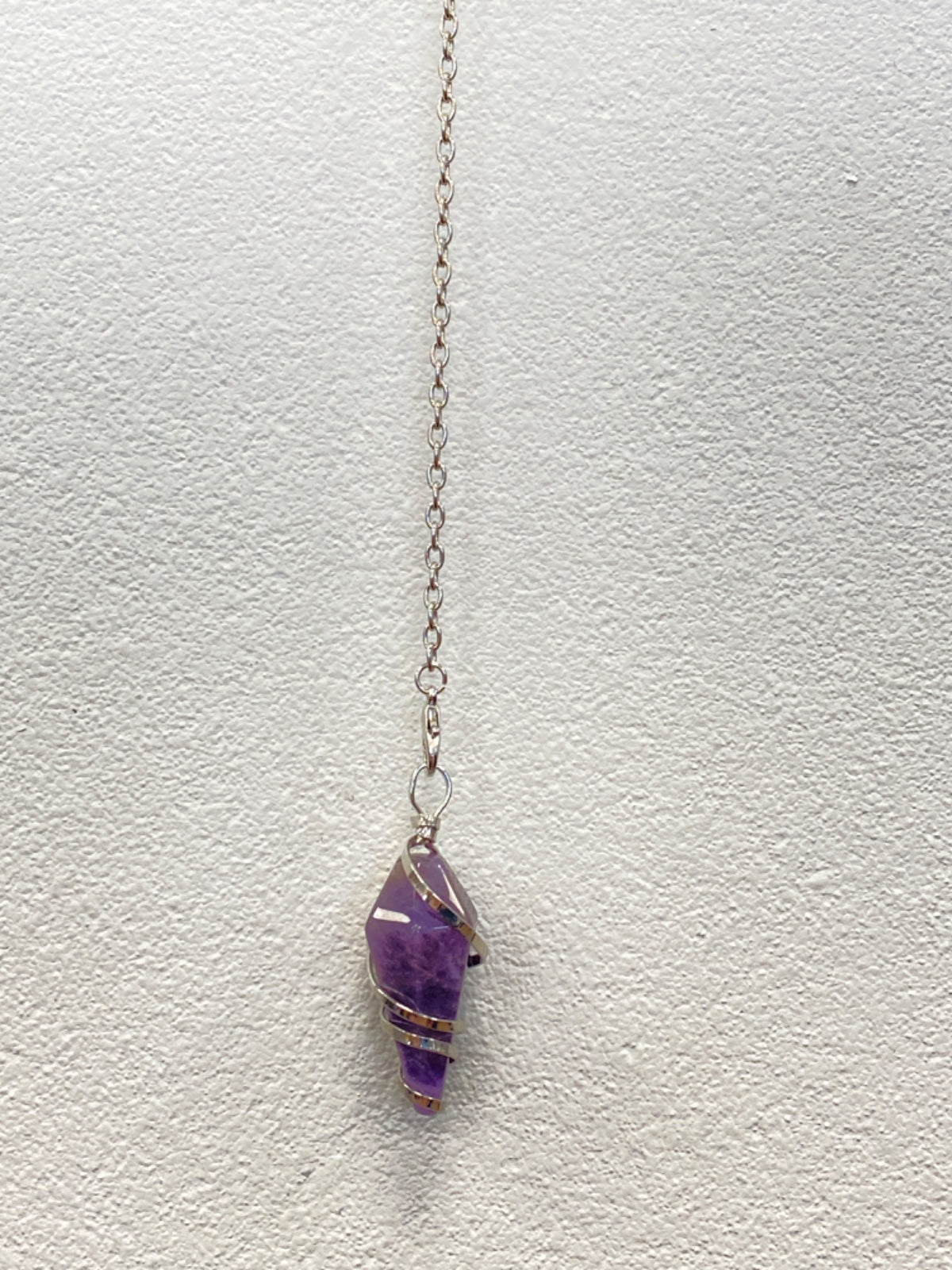 Amethyst pendulum with silver spiral wire wrap hanging from a delicate chain against a white wall